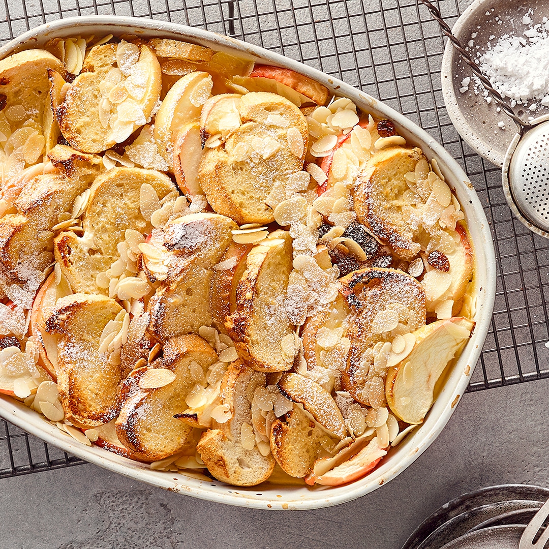 Eine Auflaufform gefüllt mit Apfel- und Brotscheiben, dazu Rosinen und Mandelblättchen, garniert mit Puderzucker