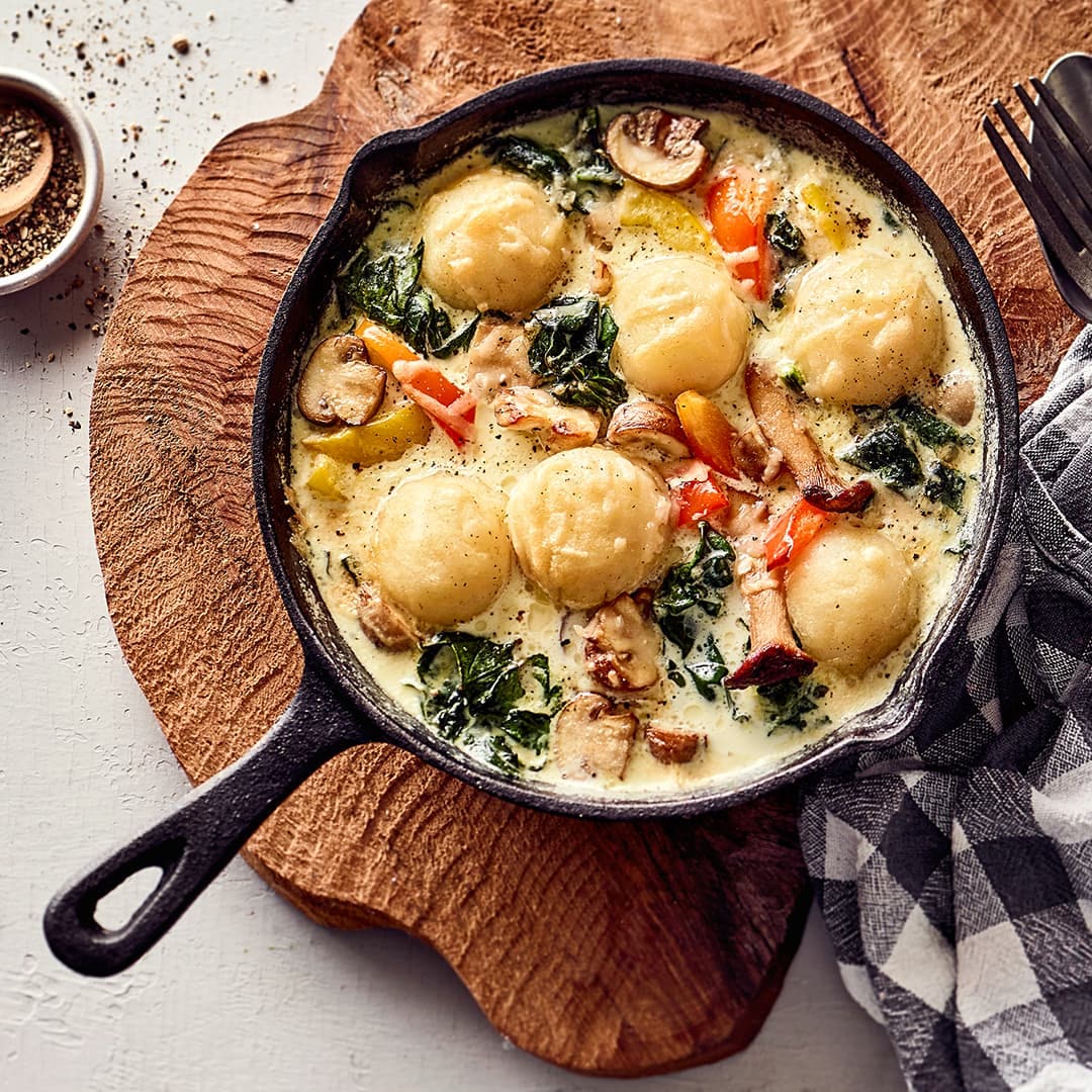 Rezept für Mini-Knödel-Pfanne Eine gusseiserne Pfanne mit hellen, runden Teigbällchen in einer hellen, cremigen Soße mit Paprika, Pizen und Spinat