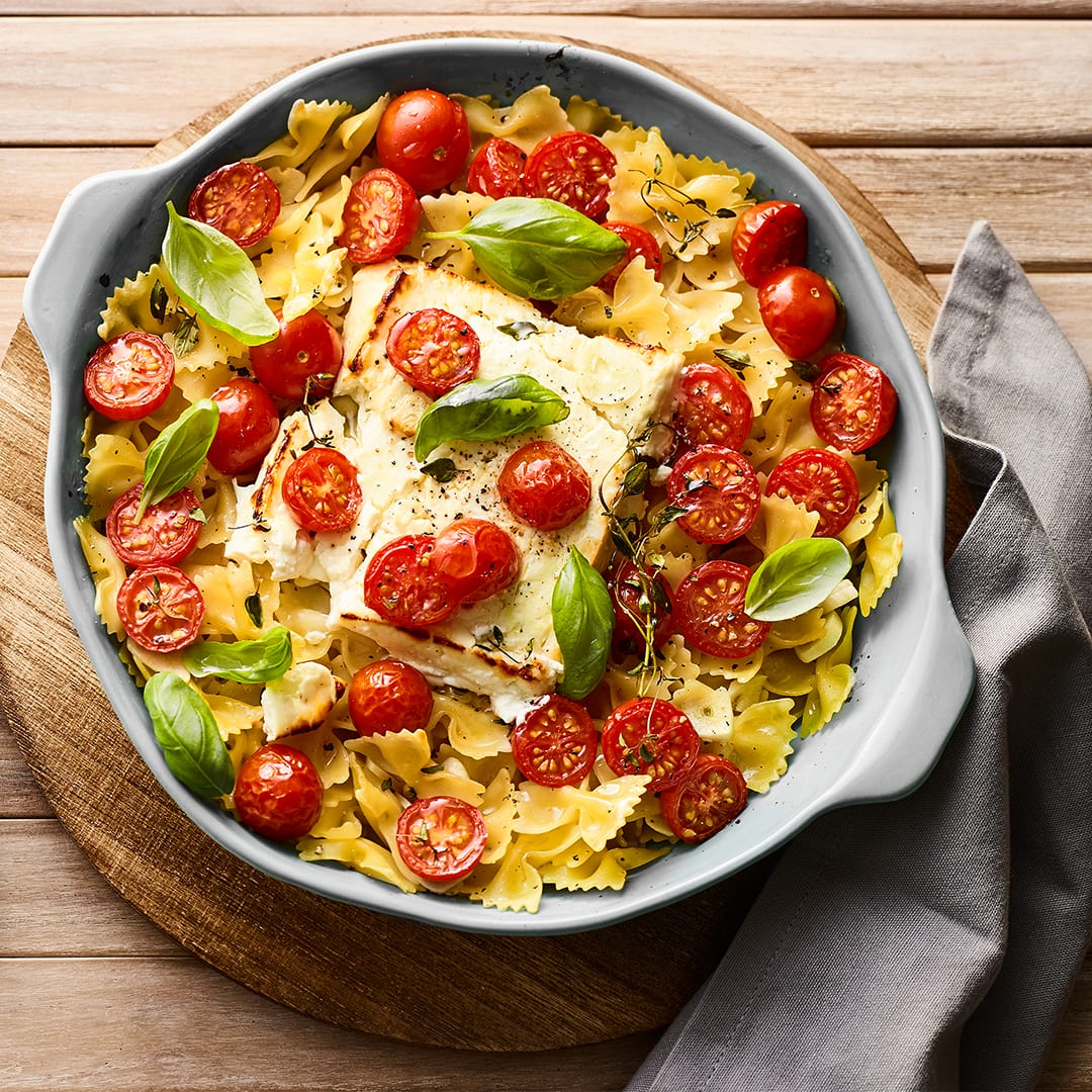 Rezept für Baked-Feta-Pasta Ein gusseiserner Topf mit Farfalle-Pasta, Kirschtomaten, einem Block gebackenem Feta und frischem Basilikum