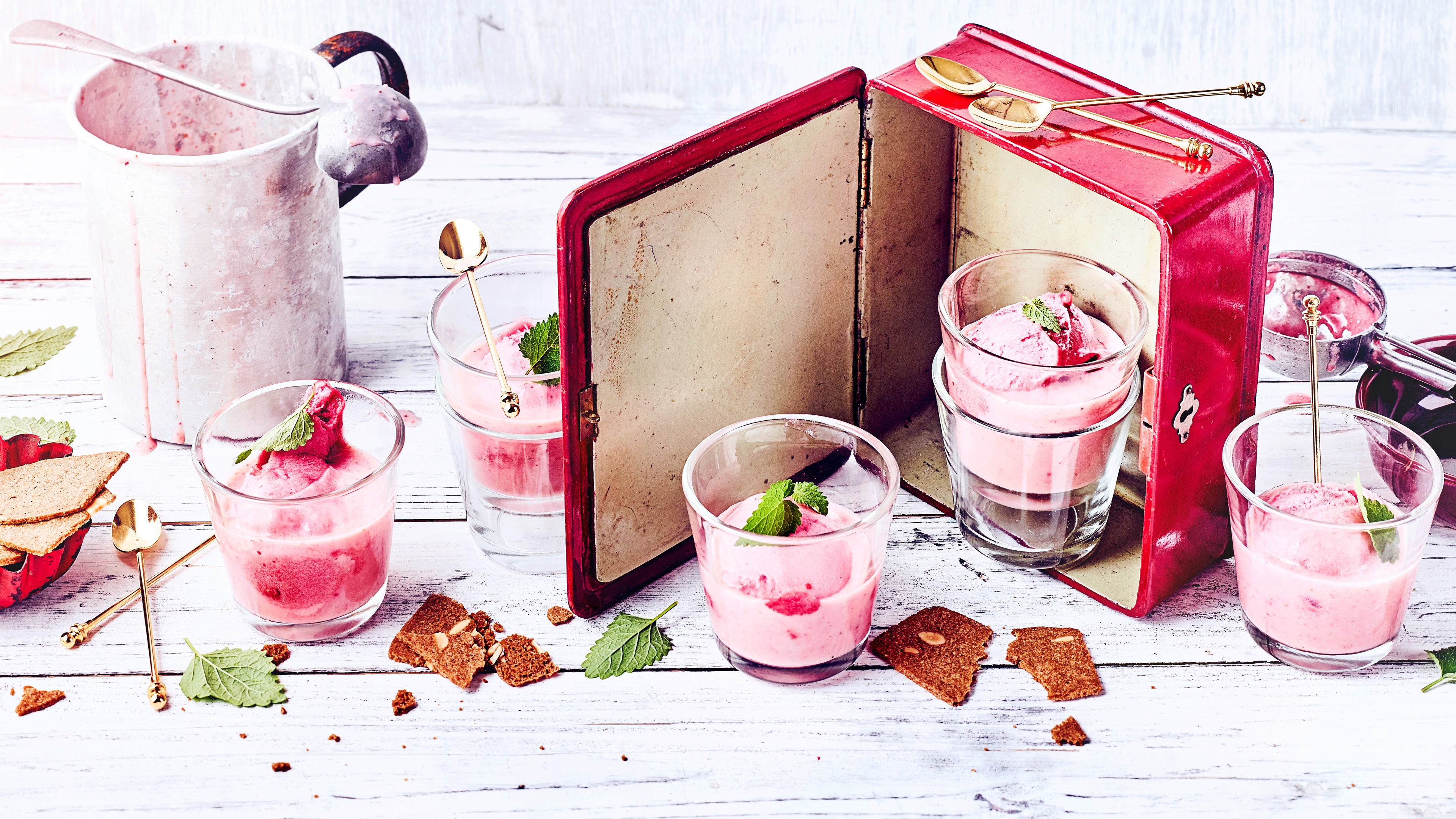 Cremiges rosa Dessert in Gläsern mit Minze auf weißem Holztisch, daneben rotes Buch und Kekskrümel Buttermilch-Erdbeer-Frappé