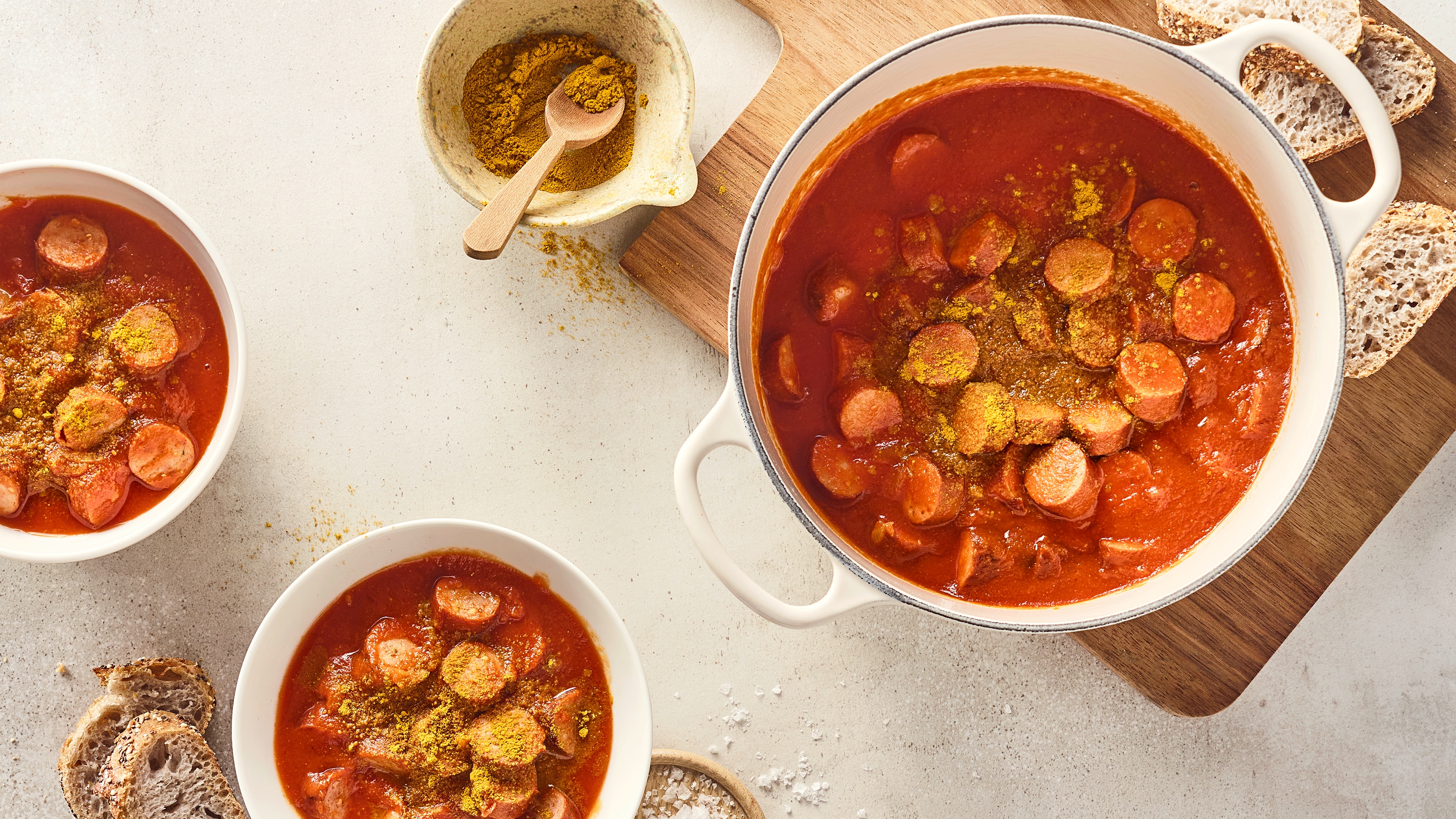 Rezept für Currywursttopf Ein gusseiserner Topf mit Wurststückchen in dunkelroter Soße, getoppt mit gelblichem Gewürz