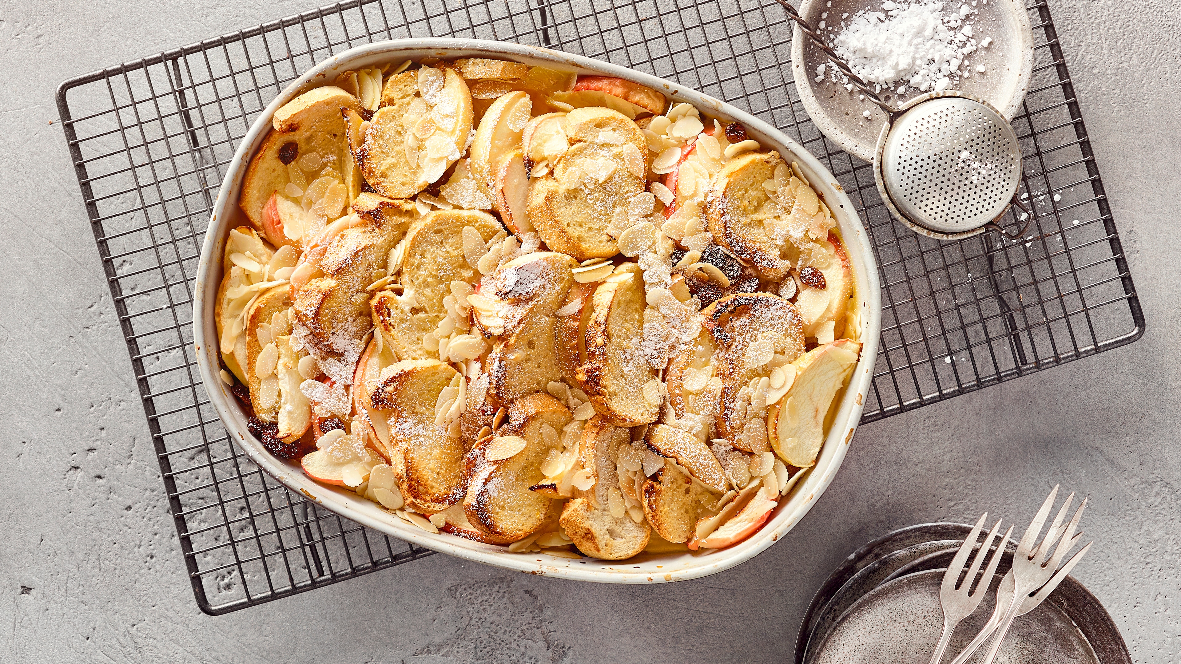 Scheiterhaufen Rezept mit Apfel und Mandel Eine Auflaufform gefüllt mit Apfel- und Brotscheiben, dazu Rosinen und Mandelblättchen, garniert mit Puderzucker